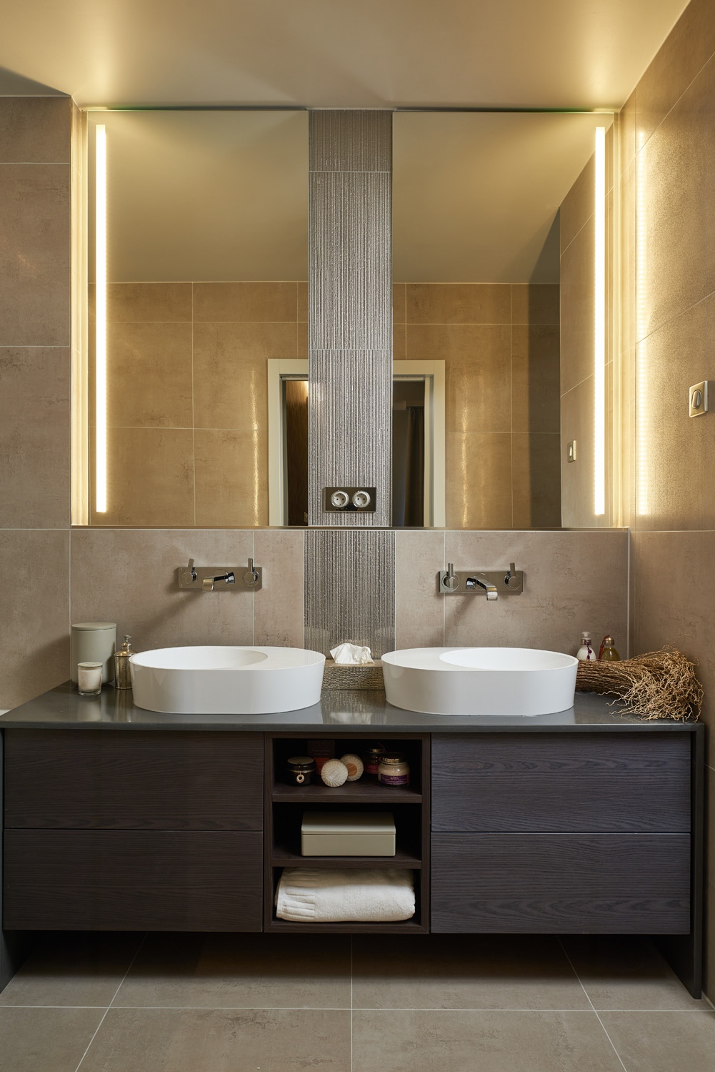 Az egyedi mosdószekrény egyenes vonalait a lekerekített mosdókagylók lágyítják a fürdőszobában, ahol a szürkés- barnás színek némi csillogással egészülnek ki