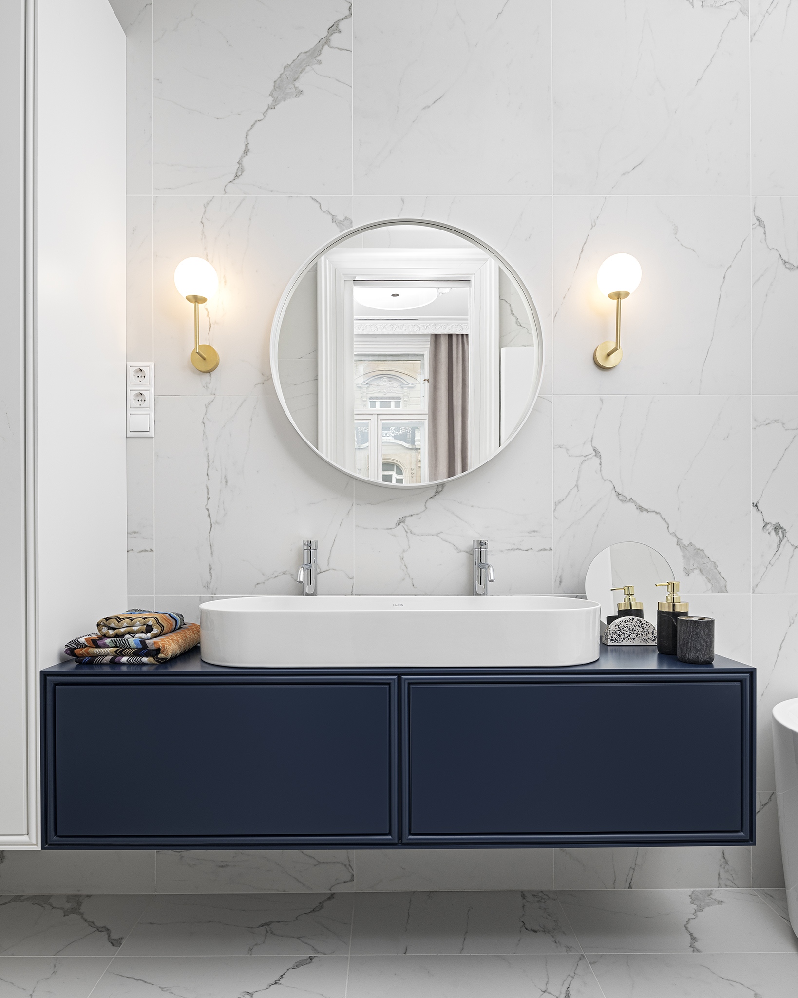 A sötétkék tónus a fürdőszoba egyedi mosdószekrényén is megjelenik – az eleganciát a márványmintás burkolat és az arany falikarok fokozzák.