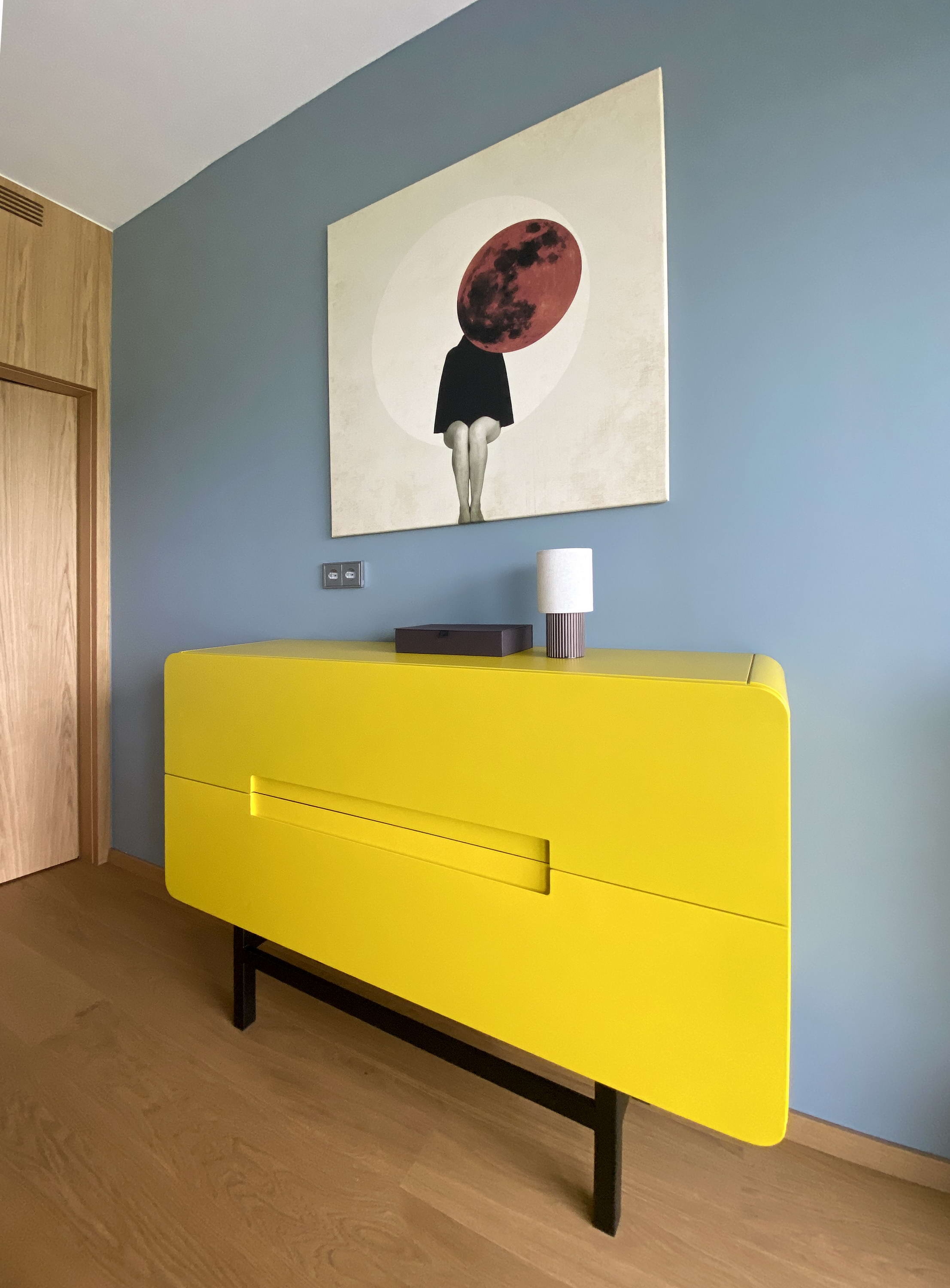 A kék háttér elé állított sárga komód a kortárs művészeti alkotással különleges színfoltot képez a hálószobában