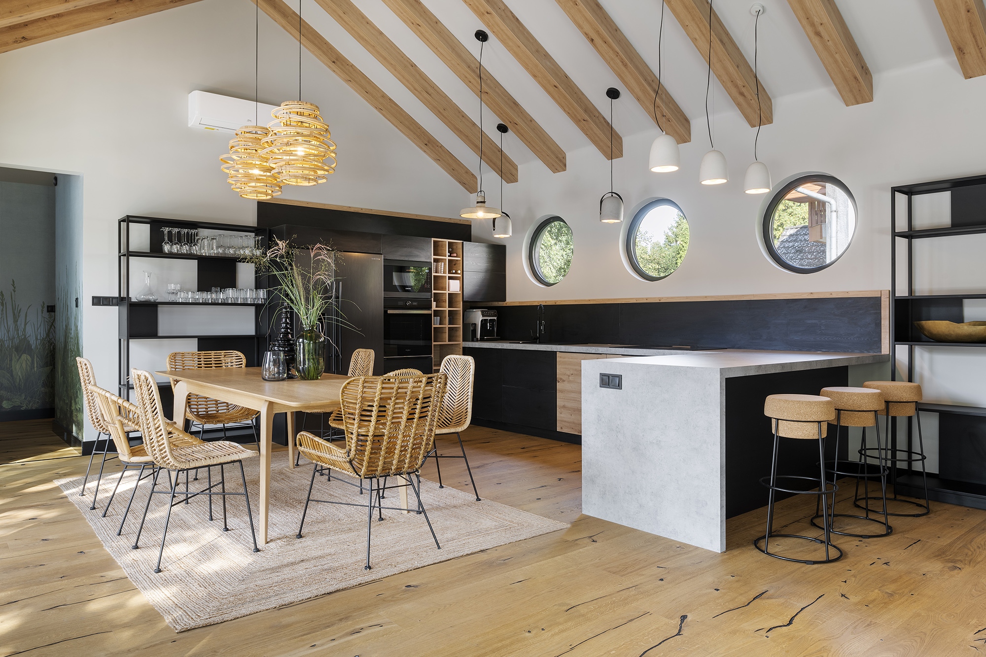  A nagyvonalú közösségi tér egyik részét a konyha és az étkező foglalja el természetes anyagokat felvonultató bútorokkal és függesztékekkel – ami hatásos kontrasztot képez a beépített bútorok fekete frontjaival és a beton konyhapulttal