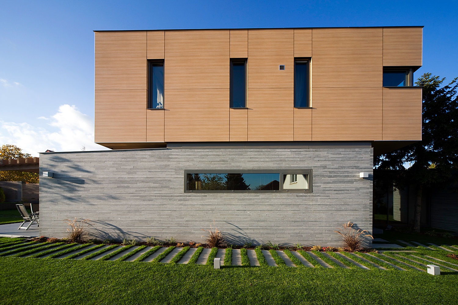 A stabil és masszív alsó szint fölött lebegni látszik az elcsúszott emelet. A légies hatást fokozza a kétféle burkolat, valamint a horizontális és vertikális osztás váltakozása